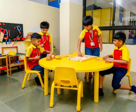 Four young students in red and yellow uniforms engage with wooden toys around a yellow table in a classroom