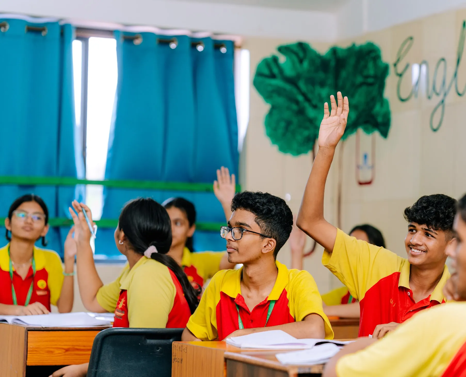 Students in yellow and red uniforms eagerly raise their hands in a classroom