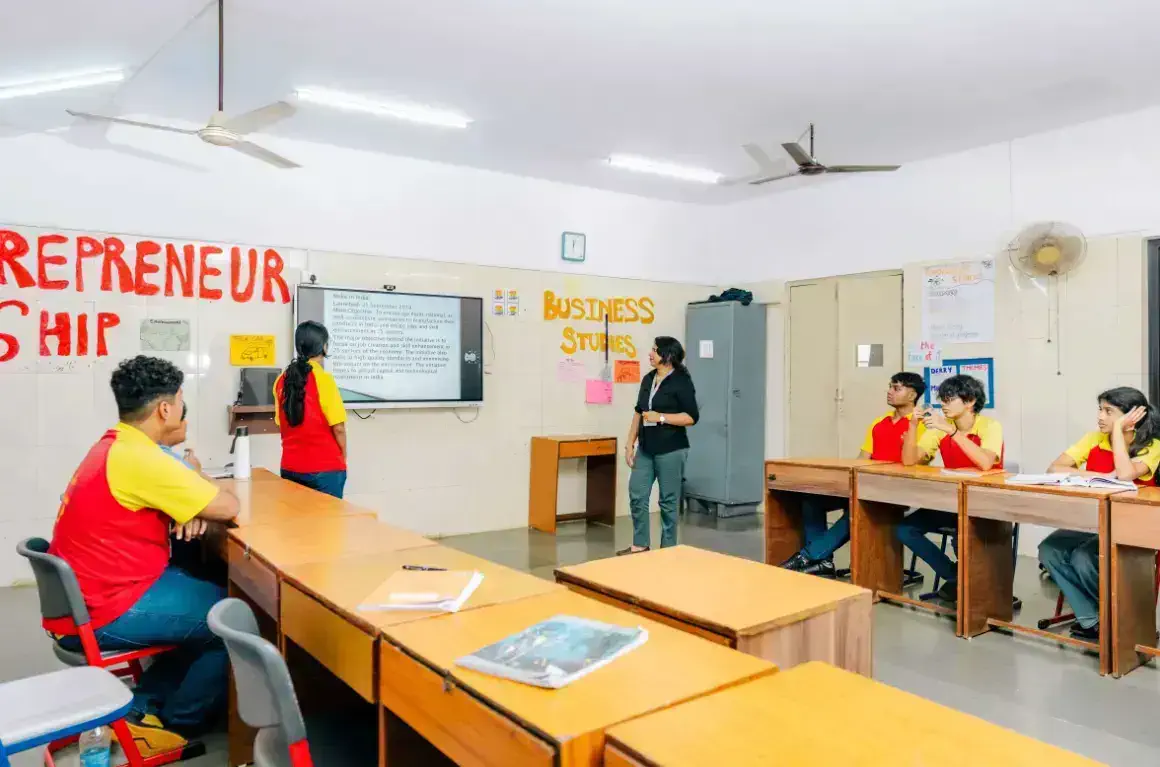 Students in a classroom face a screen with a teacher