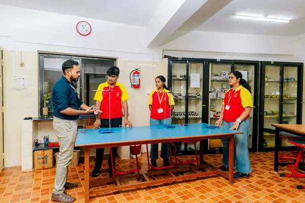A teacher explains an experiment to three students in red and yellow uniforms in a bright room