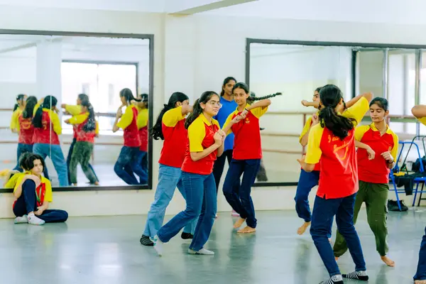 A group of young students in red and yellow uniforms dance energetically in a mirrored room with an instructor