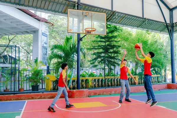 Three students play basketball on an outdoor court. The court is surrounded by green trees