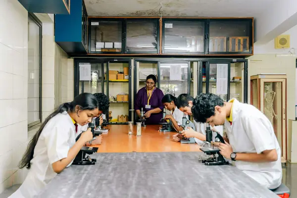 A group of students use microscopes at a lab table while a teacher observes them